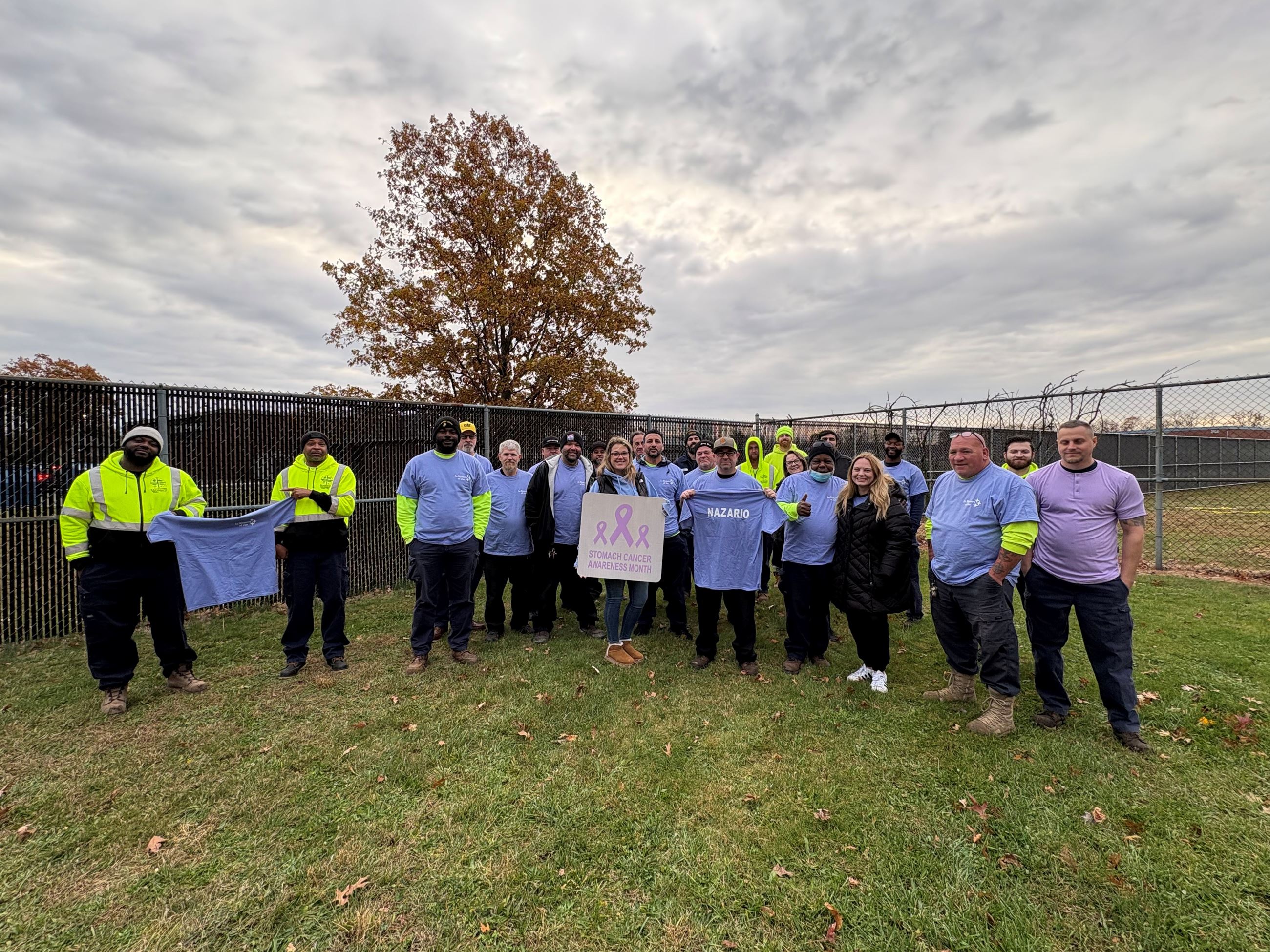 Outdoor group photo: people in neon and blue shirts hold banners reading “STOMACH CANCER AWARENESS” and “NAZARIO,” with an “&” ribbon symbol.